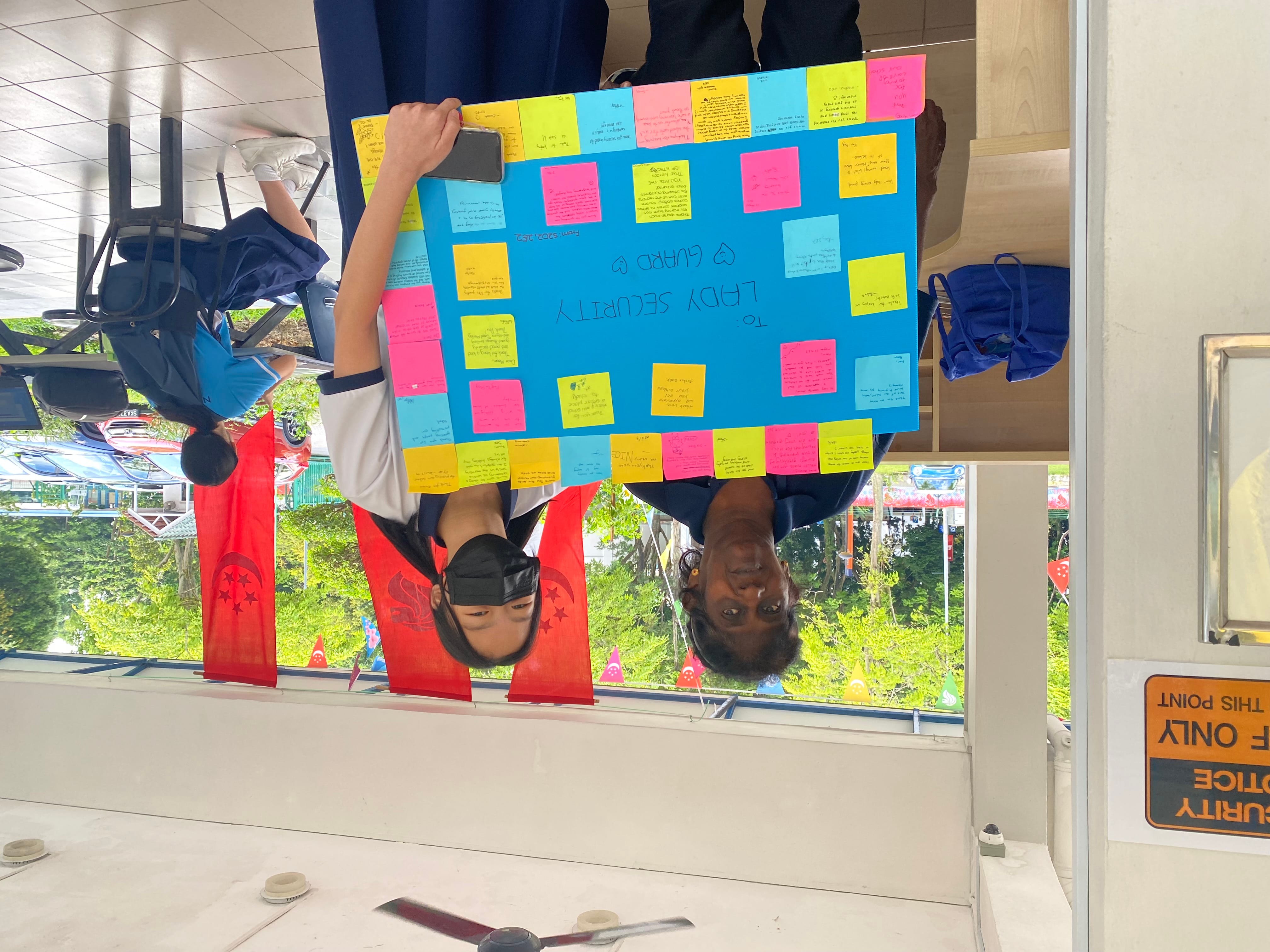 A female student passing a board of memos and post-its with messages of gratitude to the school's security guard